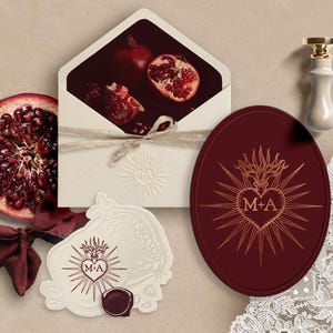 Puede incluir: Arreglo de papelería y decoración de bodas. Incluye un sobre crema con una granada, un óvalo burdeos con diseño de corazón, una tarjeta blanca en relieve, un sello de cera y una cinta.