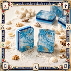 May include: Two rectangular mahjong tiles featuring a swirling blue and white marbled resin design with embedded gold flakes. The tiles have a layered blue and white side profile and are displayed alongside various small seashells and coral pieces.