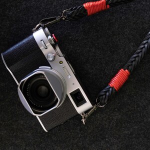 May include: A silver and black digital camera with a lens hood attached, connected to a black braided leather camera strap featuring vibrant red thread accents wrapped around the strap near the metal swivel clips on both ends.