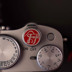 Op de afbeelding: Close-up van een zilveren camerabody met draaiknoppen en een rood-wit Fuji-logo. De camera heeft een "ON OFF"-schakelaar en diverse instellingen. De draaiknoppen zijn gemarkeerd met cijfers en symbolen.