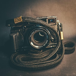 Op de afbeelding: Een zwarte digitale camera met een Fujinon Aspherical Lens Super EBC f=35mm 1:1.4 lens. De camera heeft een zwarte behuizing met textuur en is bevestigd aan een dikke, gevlochten zwarte touwriem met goudkleurige metalen accenten en een X-vormig logo.