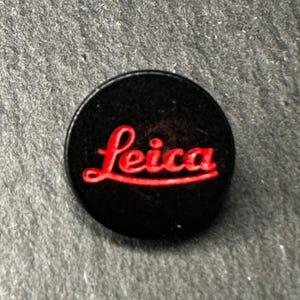 Könnte beinhalten: Ein runder schwarzer Ansteckpin mit dem roten Leica-Schriftzug in der Mitte. Das Emblem hat eine glatte, dunkle Oberfläche, auf der der Markenname in einer kräftigen, kursiven roten Schrift deutlich zu sehen ist und einen Kontrast bildet.