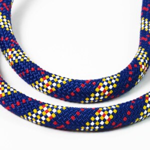 Può includere: Una corda intrecciata spessa con un colore di base blu profondo caratterizzato da un motivo a rombi ripetuto in bianco, giallo e rosso. Il cordino è flessibile e curvo, mostrando una superficie tessuta e testurizzata con distribuzione uniforme.