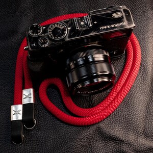 Könnte beinhalten: Eine schwarze Fujifilm X-Pro1 Digitalkamera mit Objektiv und einem leuchtend roten Kameragurt aus geflochtenem Seil. Der Gurt verfügt über silberne Metallverbinder mit einem X-Logo und schwarze Lederlaschen zur sicheren Befestigung.