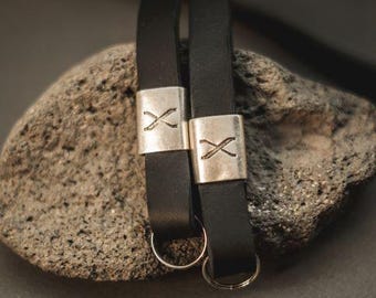 Puede incluir: Dos correas de cuero negro, cada una con una cuenta rectangular de metal plateado grabada con un símbolo X. Cada correa termina en un pequeño anillo circular de metal plateado. Las correas están colocadas verticalmente sobre una piedra texturizada.