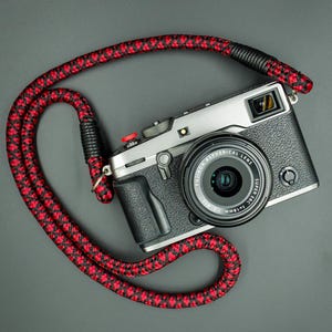 Può includere: Una fotocamera digitale argento e nera con obiettivo Fujinon Aspherical Lens Super EBC f=18mm 1:2. La fotocamera presenta un pulsante di scatto rosso ed è collegata a una tracolla in corda intrecciata con motivo geometrico nero e rosso.