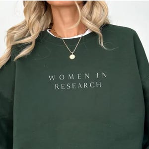 Puede incluir: Una sudadera verde oscuro con el texto "WOMEN IN RESEARCH" en blanco. La sudadera tiene cuello redondo y mangas largas. Se ve una camiseta blanca debajo. La modelo lleva pantalones cortos negros y un collar dorado.