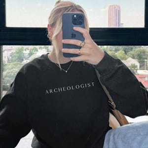 Peut inclure: Sweat-shirt noir avec le mot "ARCHEOLOGISTE" imprimé en blanc. Le sweat-shirt est porté avec un jean bleu clair. Une personne prend un selfie, tenant un téléphone devant son visage.