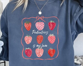 May include: A blue-grey sweatshirt featuring a strawberry design with the text "Pediatrics is my jam." The strawberries are red and pink, arranged in a square with a pink border. The sweatshirt has a relaxed fit.