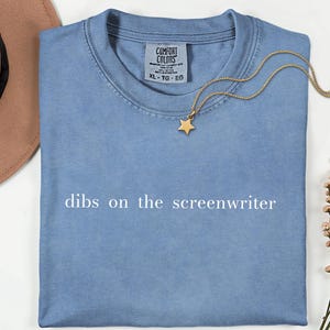 May include: A blue short-sleeve t-shirt with the text "dibs on the screenwriter" printed in white across the chest. The shirt features a Comfort Colors brand label at the inner neckline, indicating an XL size, and is made of 100% cotton fabric.