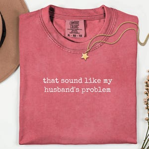 May include: A dusty rose crewneck t-shirt featuring the white text "that sound like my husband's problem" printed on the chest. The shirt has a Comfort Colors label at the inner neckline, a gold star pendant necklace, a tan hat, and dried flowers.