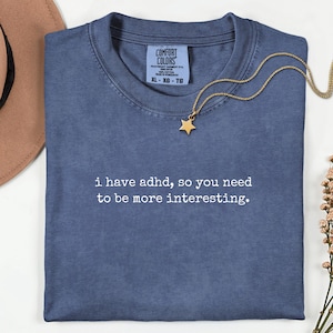 May include: A blue short-sleeved t-shirt with the white text "i have adhd, so you need to be more interesting." printed on the front. The shirt features a Comfort Colors label in the neck area, a gold star pendant necklace, a tan hat, and dried flowers.