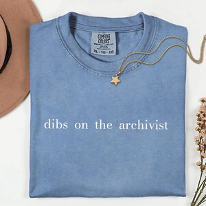 May include: A blue t-shirt with the text "dibs on the archivist" printed in white across the chest. The shirt features a Comfort Colors brand label at the neckline. A gold-toned necklace with a small star pendant rests on the fabric.
