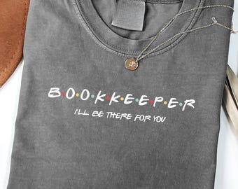 May include: A gray t-shirt with the word "BOOKKEEPER" in a colorful font, and the phrase "I'LL BE THERE FOR YOU" below it. A gold necklace with a pendant is on the shirt. A brown hat and decorative leaves are in the background.