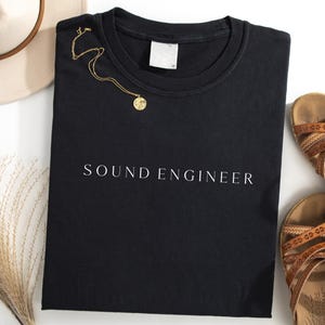 Może przedstawiać: Czarna bluza z okrągłym dekoltem z białym napisem "SOUND ENGINEER". Złoty naszyjnik z wisiorkiem jest ułożony na koszulce. Beżowy kapelusz i brązowe sandały również znajdują się na zdjęciu.