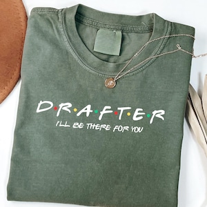 Puede incluir: Una camiseta verde oliva con la palabra "DRAFTER" en letras blancas, con las letras "A", "F" y "E" en rojo, amarillo y verde. Debajo está la frase "I'LL BE THERE FOR YOU". Un collar dorado con un colgante está sobre la camiseta.