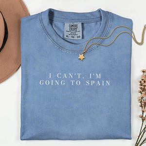 May include: A blue t-shirt with the white text I CAN'T, I'M GOING TO SPAIN printed on the front. The shirt features a Comfort Colors label at the neckline, size XL, and is accompanied by a gold-toned necklace with a small star-shaped pendant.
