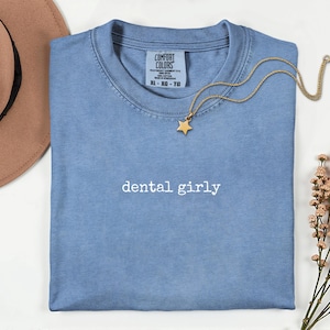 以下が含まれることがあります： 胸元に白い刺繍でdental girlyと書かれた青い半袖Tシャツ。首元にはComfort Colorsのラベルがあり、サイズはXL-XG-TGと記載されている。生地の上にはゴールドの星型ペンダントネックレスが置かれている。