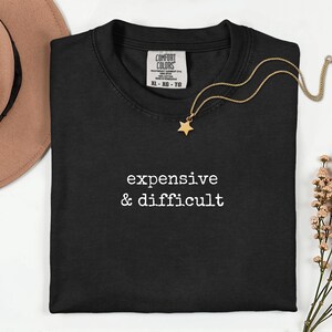 Può includere: Una t-shirt nera a maniche corte con il testo bianco "expensive & difficult" stampato sul petto. La maglietta presenta un'etichetta del marchio Comfort Colors sul collo che indica la taglia XL. Una collana color oro con ciondolo a stella.