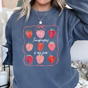 Puede incluir: Sudadera azul grisáceo con diseño de fresas y el texto "Sonography is my jam". El diseño incluye un marco rosa con nueve fresas en varios tonos de rojo y rosa. La sudadera tiene mangas largas.