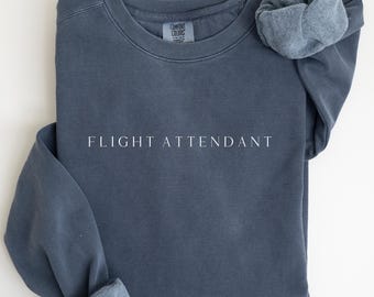 May include: A slate blue crew neck sweatshirt with the words "FLIGHT ATTENDANT" printed in white. The sweatshirt has rolled-up sleeves and a Comfort Colors label.