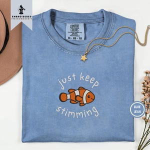 May include: A blue jean colored t-shirt featuring an embroidered orange and white clownfish graphic with the text just keep stimming. The shirt has a crew neckline and a Comfort Colors label at the inner collar. A gold star pendant necklace is included.