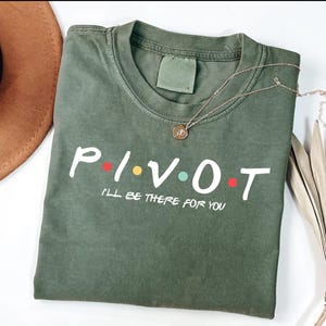 Könnte beinhalten: Olivgrünes T-Shirt mit dem Wort "PIVOT" in Weiß, mit bunten Punkten. Unter dem Wort "PIVOT" steht der Text "I'LL BE THERE FOR YOU". Eine goldene Halskette mit Anhänger liegt auf dem Shirt.