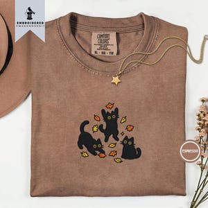 Può includere: Una t-shirt marrone con un ricamo raffigurante tre gatti neri che giocano con foglie autunnali arancioni e gialle. La maglietta presenta un'etichetta Comfort Colors, un girocollo ed è abbinata a una collana con ciondolo a forma di stella.