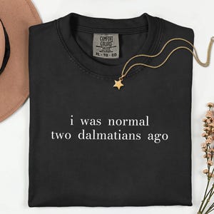 May include: A black short-sleeved t-shirt with the white text "i was normal two dalmatians ago" printed on the front. The shirt features a Comfort Colors brand label at the inner neckline, indicating an XL size, and a crew neck collar design.