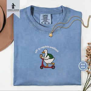 Könnte beinhalten: Ein blaues T-Shirt mit einer Stickerei, die eine weiße Ente mit grünem Rucksack auf einem roten Roller zeigt, die einen Kaffeebecher hält. Der Text über der Ente lautet off to regulate emotions in weißen Buchstaben.