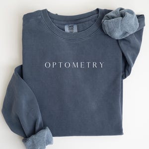 May include: A slate blue sweatshirt with the word "OPTOMETRY" printed in white. The long-sleeved shirt has a crew neck and a soft, fleece-lined interior. The sleeves are cuffed, and the shirt is laid flat on a white surface.