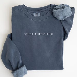 May include: A slate blue sweatshirt with the word "SONOGRAPHER" printed in white. The long-sleeved shirt has folded cuffs and a crew neck. The Comfort Colors brand tag is visible at the top.