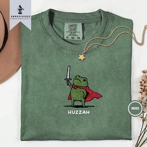Pode incluir: Uma t-shirt de gola redonda em verde musgo com um bordado de um sapo verde a usar uma capa vermelha e a segurar uma espada prateada. Abaixo do sapo, a palavra HUZZAH está impressa em branco. A t-shirt tem uma etiqueta Comfort Colors no pescoço.