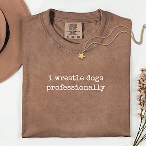 Puede incluir: Una camiseta marrón con el texto blanco "i wrestle dogs professionally" impreso en el frente. La prenda incluye una etiqueta de la marca Comfort Colors en el cuello, un sombrero beige de ala ancha, un collar con estrella dorada y flores.