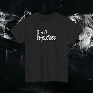 May include: Black crew neck t-shirt with the word "lifelover" in white script across the chest. The shirt is a solid colour and has short sleeves. The background is a dark, abstract design.