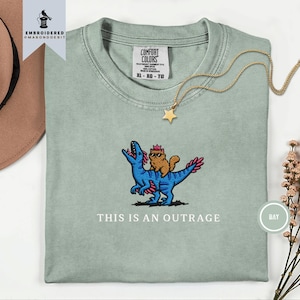 May include: A sage green t-shirt featuring an embroidered graphic of a blue dinosaur with pink accents carrying a brown cat wearing sunglasses and a pink crown. The text "THIS IS AN OUTRAGE" is embroidered below the graphic in white lettering.