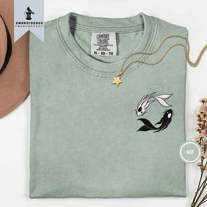 Könnte beinhalten: Ein salbeigrünes T-Shirt mit Rundhalsausschnitt und einer Stickerei eines Yin-Yang-Motivs aus zwei Koi-Fischen, einem weißen und einem schwarzen. Das T-Shirt hat ein Comfort Colors-Etikett im Nacken, Größe XL, und liegt neben einer goldenen Sternkette.