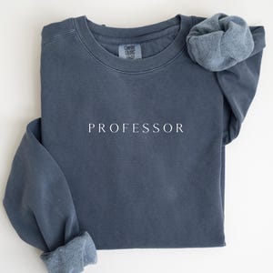 Puede incluir: Sudadera azul pizarra con la palabra "PROFESOR" impresa en letras blancas. La sudadera tiene cuello redondo y mangas largas con puños doblados. La etiqueta de la marca "COMFORT COLORS" es visible.