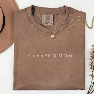 Könnte beinhalten: Ein braunes T-Shirt mit dem weißen Text "CAVAPOO MOM". Eine goldene Sternanhänger-Halskette und ein brauner Hut sind ebenfalls zu sehen. Das Shirt hat ein Comfort Colors-Etikett.