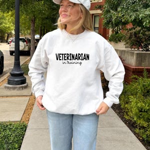 May include: A white sweatshirt with the text "VETERINARIAN in training" in black. The sweatshirt is long-sleeved and worn with light blue jeans and a white cap. The person is standing outside on a sidewalk.