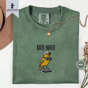 Op de afbeelding: Een mosgroen t-shirt met een geborduurde afbeelding van een gele vogel die een grijze honkbalpet en grijze laarzen draagt en een verrekijker vasthoudt. De tekst BIRD NERD is boven de vogel geborduurd. Het shirt heeft een Comfort Colors label.