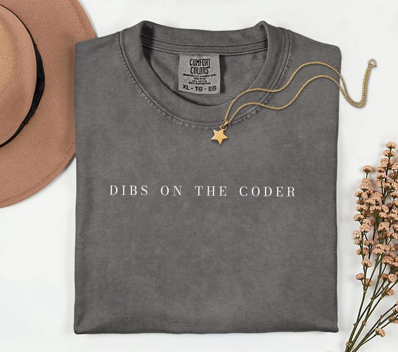 May include: A charcoal grey crew neck t-shirt featuring the white text DIBS ON THE CODER printed across the chest. The shirt has a Comfort Colors brand label at the inner neckline and is displayed with a gold star pendant necklace.