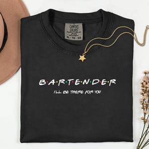 Può includere: T-shirt nero girocollo con la scritta "BARTENDER" in un font colorato e la frase "I'll be there for you". Una collana d'oro con un ciondolo a forma di stella è sulla maglietta.