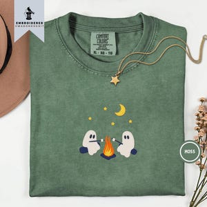 May include: A moss green short-sleeve t-shirt featuring an embroidered graphic of two white ghosts sitting by a campfire roasting marshmallows under a yellow crescent moon and stars. The shirt has a Comfort Colors label in the neck area.