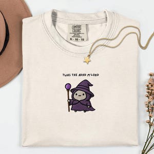 Könnte beinhalten: Ein cremefarbenes T-Shirt mit einer Stickerei, die eine kleine Figur in einem violetten Zaubererhut und Umhang zeigt, die einen Stab mit einer violetten Kugel hält. Über der Figur steht der Text TWAS THE ADHD M'LORD in schwarzen Buchstaben.