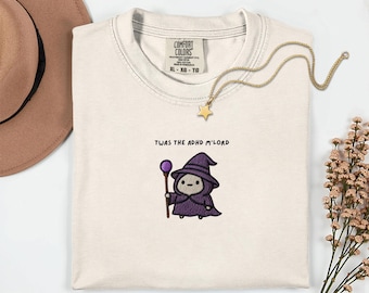 May include: A cream-colored t-shirt featuring an embroidered graphic of a small character wearing a purple wizard hat and robe, holding a staff with a purple orb. Above the character, the text reads TWAS THE ADHD M'LORD in black letters.