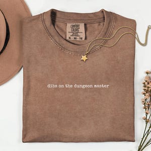 May include: A brown short-sleeved t-shirt with the text "dibs on the dungeon master" printed in white. The shirt features a Comfort Colors label indicating size XL, a gold-toned star pendant necklace, a tan wide-brimmed hat, and dried flowers.
