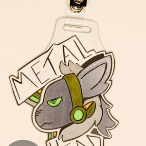 May include: A badge with a cartoon wolf wearing headphones, the words "METAL HEAD" are written above and below the wolf. The badge has a clear plastic holder with a metal clip at the top. The wolf is gray with green eyes and a green and gold headset.
