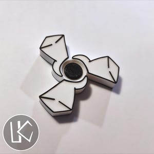 May include: A three-pronged fidget spinner featuring a white body with bold black outlines and a textured black circular centre cap. The design has a geometric, angular aesthetic with sharp edges on each of the three symmetrical arms.