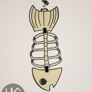 May include: A decorative fish skeleton-shaped tag in cream and black. The tag is made of layered material and has a black ribbon for attachment. A small metal clip is attached at the top.
